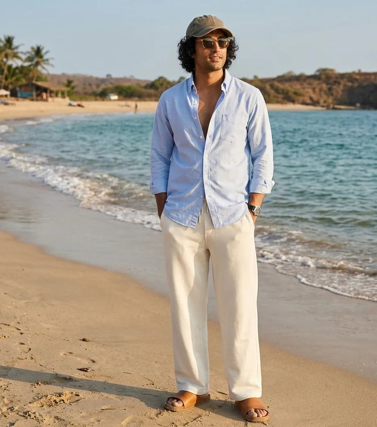 Man standing on a beach wearing a light blue shirt and beige pants with sunglasses and a cap.